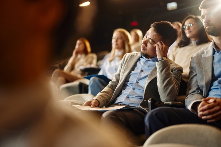 A man leans on his hand and takes a nap in a seminar room while surrounded by other awake people