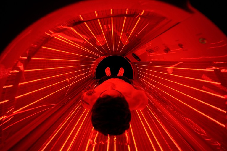 A person lies in a red-light therapy bed that entirely surrounds him with long red tube lamps.