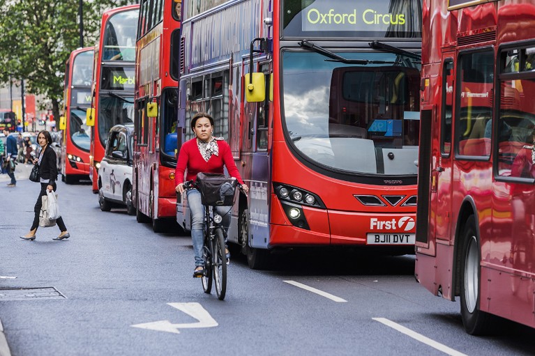 A woman rides a bike in the street alongside several London busses in the bus lane while pedestrians cross the street behind her.