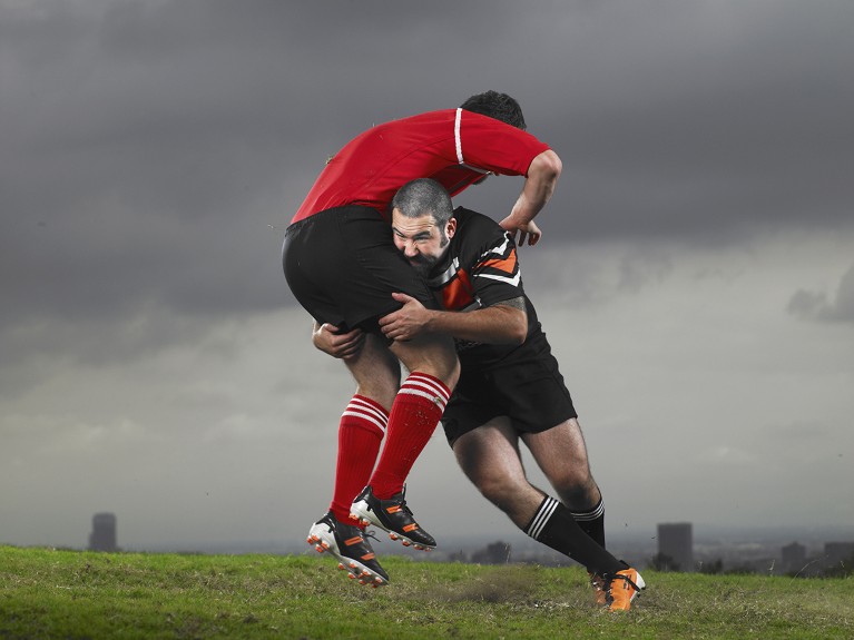 Two men from opposing teams tackle while playing rugby on a field of grass with grey clouds above.