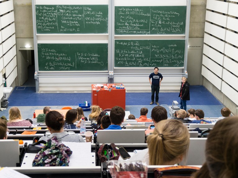 Chalkboards with scientific formulas in the background of a lecture hall filled with university students.