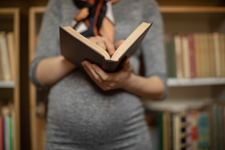 Close up view of an anonymous pregnant woman holding a book in front of library bookshelves.