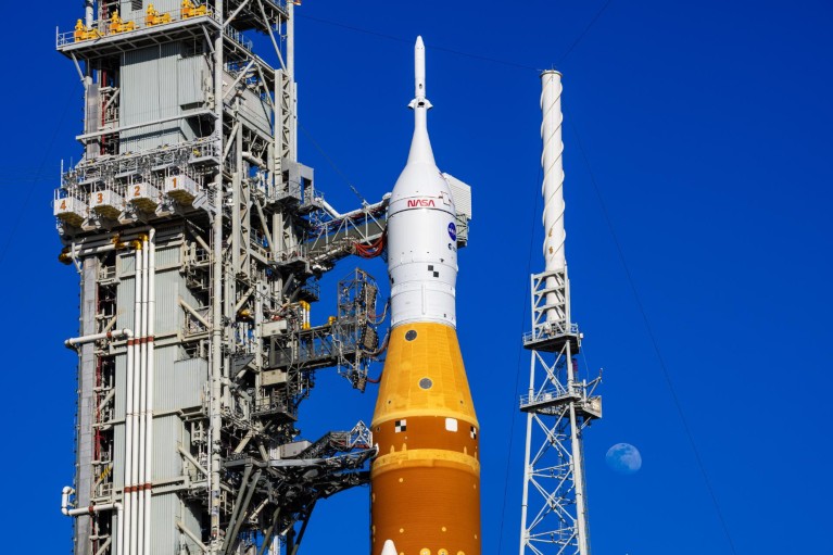 A blue sky and moon seen behind a huge rocket with the Orion spacecraft on top.