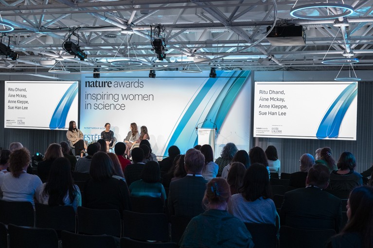 A panel of four women sit in a darkened auditorium full of people. Bright screens are illuminate the words “Nature Awards; Inspiring Women in Science”.