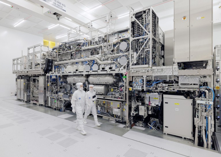 Two technicians in full white suits and face masks walk in front of a large EUV lithography system inside a bright laboratory