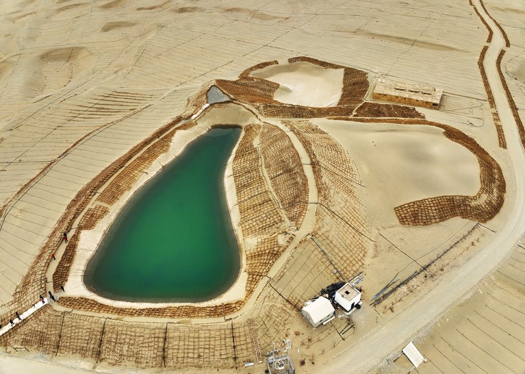 Aerial view of green lake surrounded by an embankment covered in a grid pattern, in a sandy landscape.