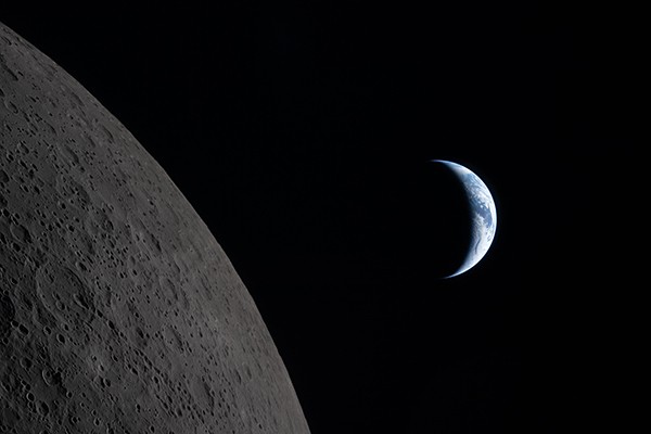 Earth, partly shadowed by the Moon and bedecked with swirling clouds, is seen beyond the pockmarked surface of the Moon.