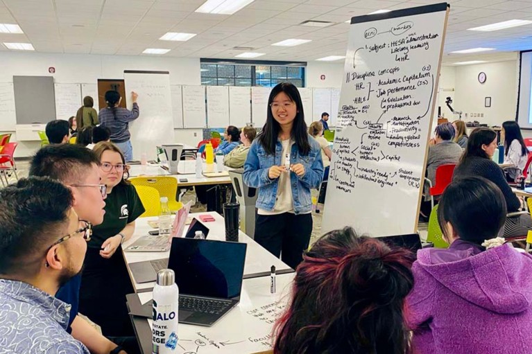 Standing beside a whiteboard of notes, Yulu Hou (center) engages with peers during a teaching and learning workshop.