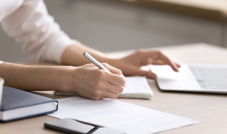 Close up female hands holding a pen writing in note book while her other hand scrolls on a laptop computer.