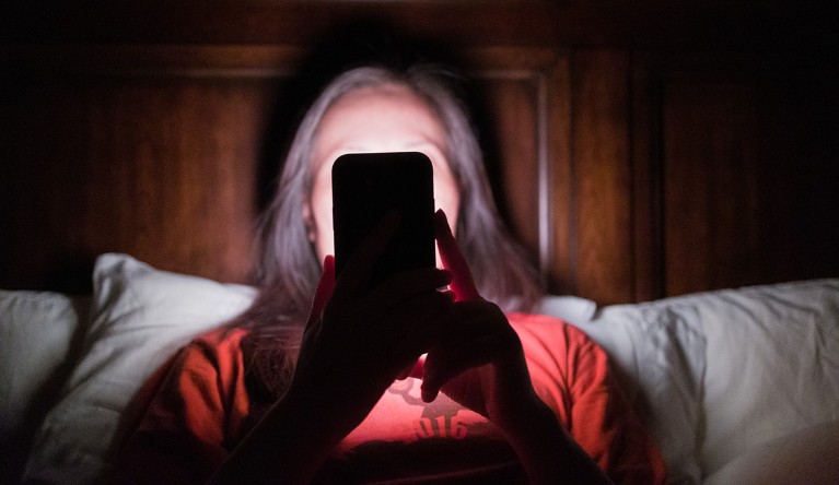 A woman laying in bed holds up a smartphone in front of her face. Her face and bed are illuminated by bright white light emitted from the phone display.