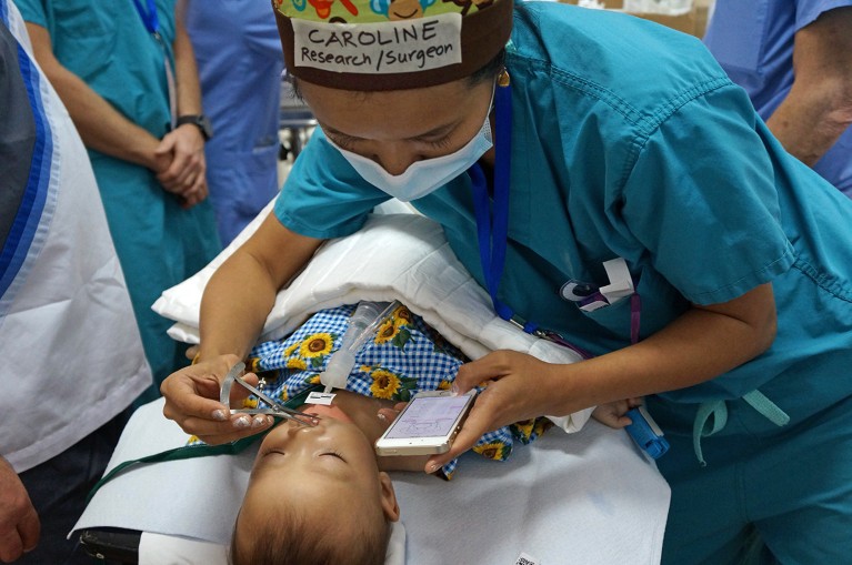 Caroline Yao wearing blue scrubs, a face covering, and a head covering with her name and job title on the front, is seen checking the cleft lip of a child in a hospital. She is holding calipers in one hand as she cross references a mouth chart on her mobile phone, held in her other hand.