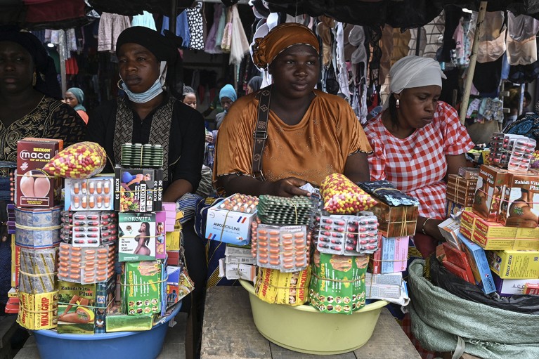 Three women sit in a row, each holding a bucket containing packets of capsules. Behind them clothes and underwear can be seen hanging.