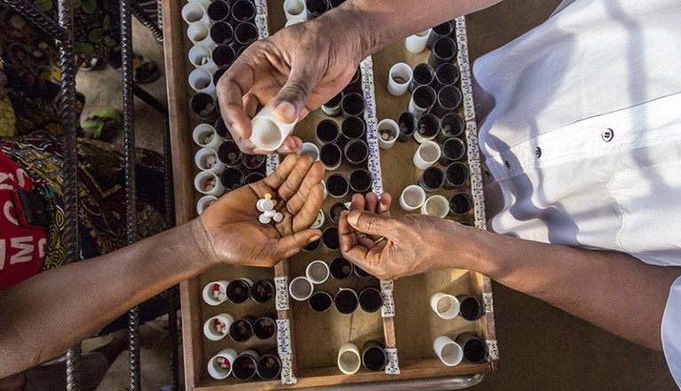 View from above shows the lower body, arm and open hand of someone holding several pills. Opposite the torso, two arms and hands of someone are seen holding a small container. Beneath the hands is a tray with containers of pills.