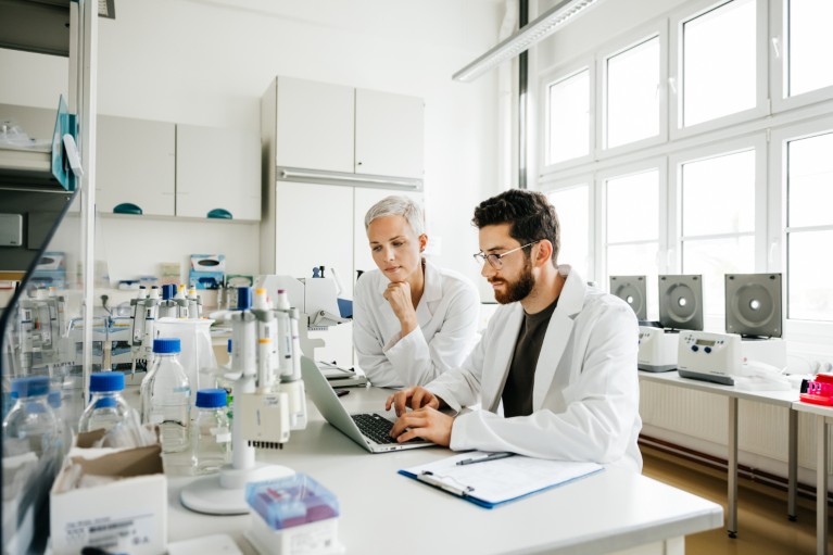 Two lab workers sat at a lab bench, male colleague working on laptop and female worker leaning in.