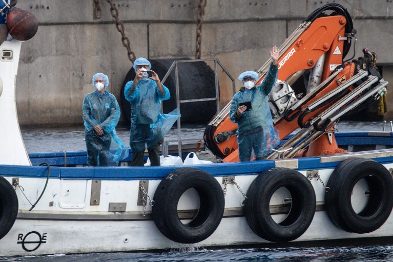 Rescued passengers from the MV Hondius, seen wearing PPE, standing and waving from a fishing boat.