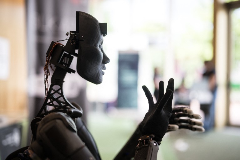 Side-profile view of a humanoid robot with exposed wiring behind its face, articulating with its hands as if speaking