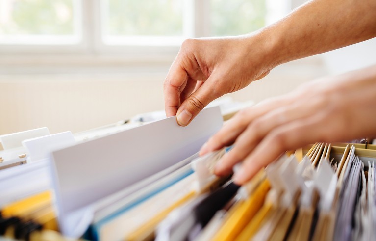 Hands sorting through a filing cabinet with multiple coloured folders, primarily white and yellow.
