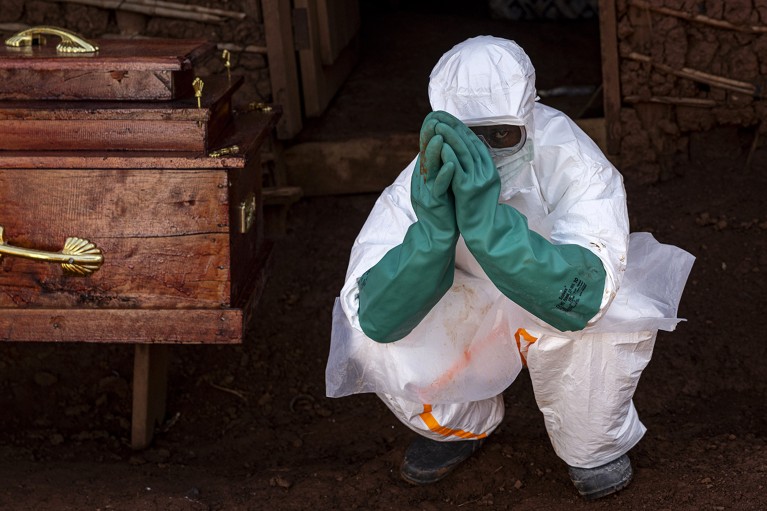 A health worker wearing protective equipment crouches beside the coffin of a suspected Ebola victim.