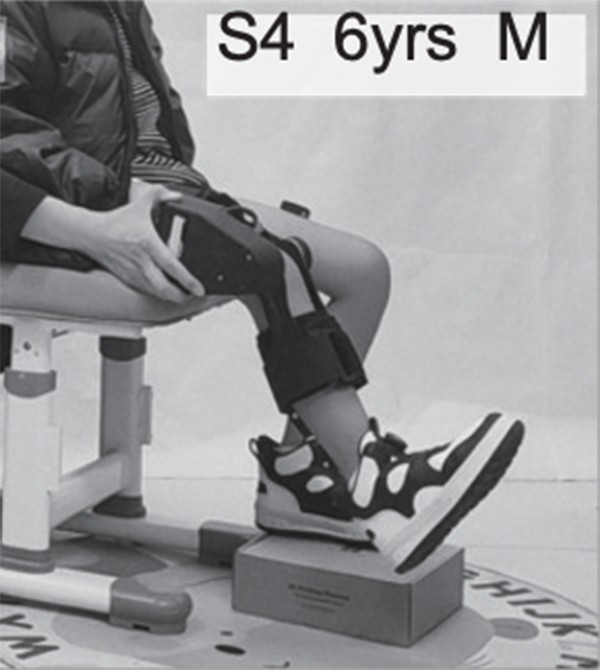 A child sitting on a chair extends one of his legs with the help of a robotic device worn around it.