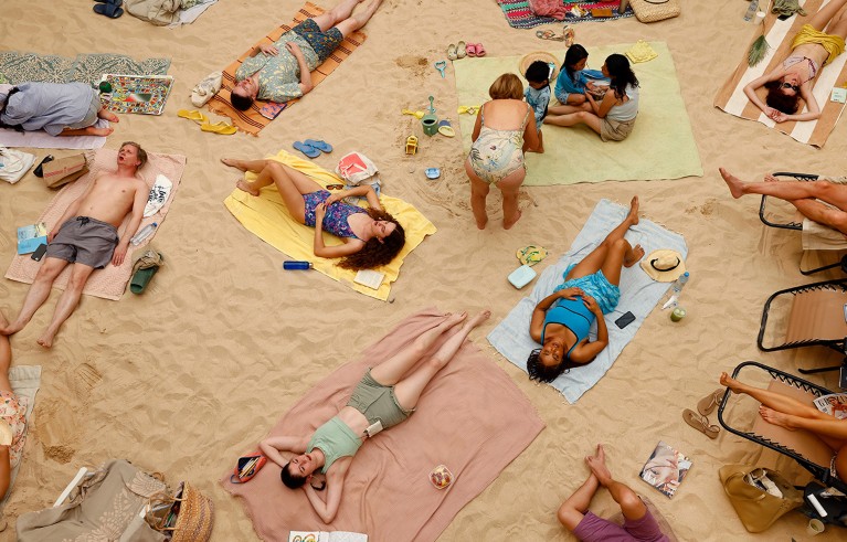 Crowd of people relaxing on towels spread across sandy beach, engaging in sunbathing and casual activities. Brightly coloured towels and scattered personal items like hats, books, and drinks.