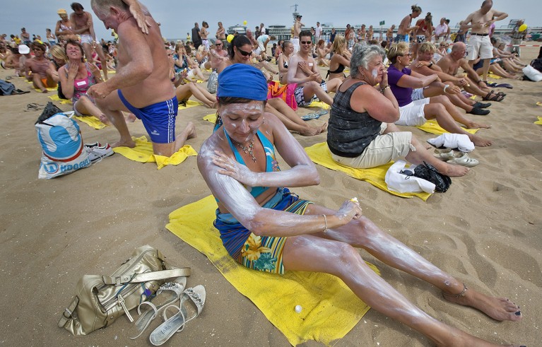 A crowded beach scene showing numerous people sitting or lying on yellow towels on sand, many applying sunscreen. Central focus on a person in a blue headscarf and patterned swimsuit, covered in white sunscreen lotion.