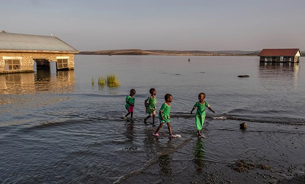 A group of four school children walk through the shallows past submerged and abandoned school buildings.