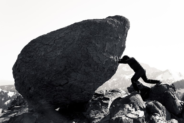 A black and white photograph of a woman futilely pushing huge boulder.