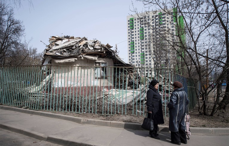 Two people standing near a metal fence around a partially collapsed building with rubble on its roof and on the ground. A tall residential building with green and white panels is visible in the background.