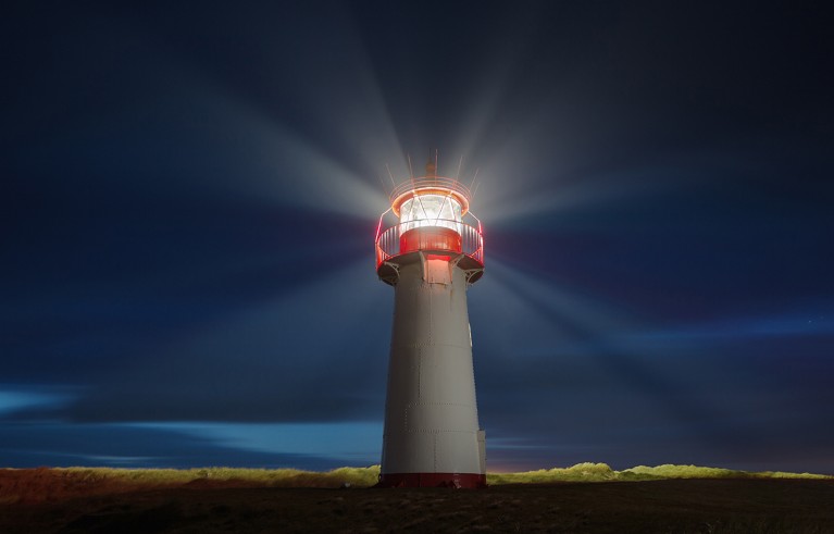 Lighthouse at night emitting bright white and red beams of light in multiple directions against a dark blue sky. The lighthouse stands on a dark, grassy terrain with a red base and a glowing red light enclosure at the top.