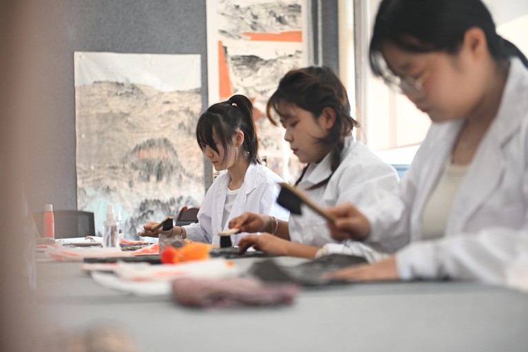 Three lab-coated students take part in a cultural relic restoration class at Lanzhou Vocational Technical College, with artwork displayed on the wall.