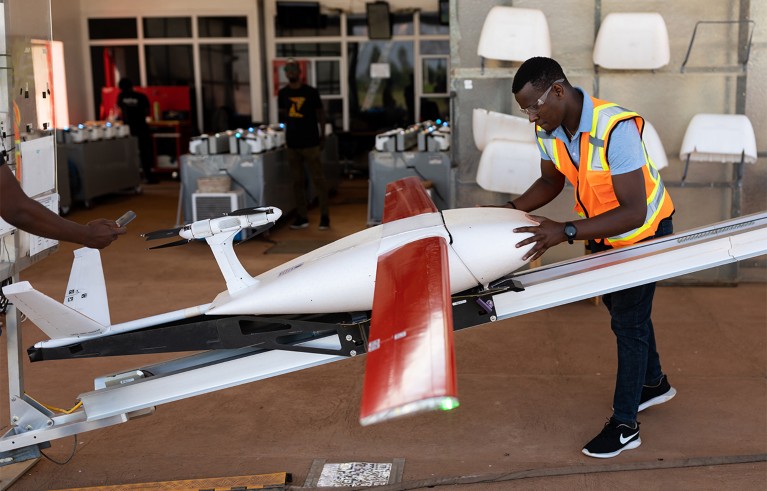 A black young man in a high-visibility vest inspecting a large drone with red and white wings on a metal stand.