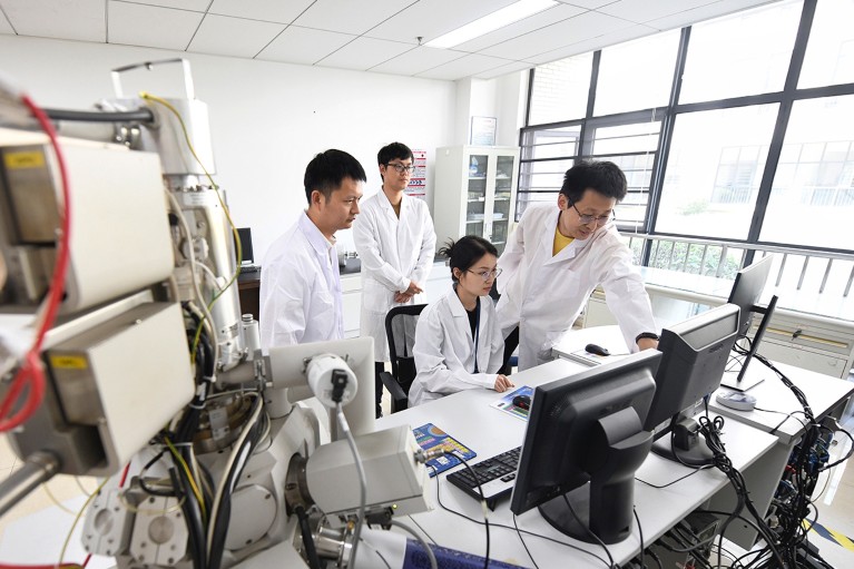 Three young Chinese scientists in lab coats, looking at a computer monitor, one of them is seated and there is a fourth older scientists offering advice.