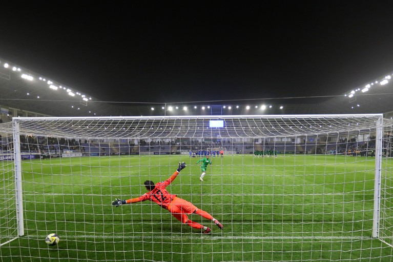 A rear view from behind the goal captures the goalkeeper diving in an attempt to save a penalty, as Sierra Leone scores against Azerbaijan during the shootout.