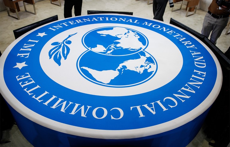 A large circular table with the International Monetary Fund (IMF) logo prominently displayed in blue and white at a meeting venue. The logo features a world map and laurel branches.