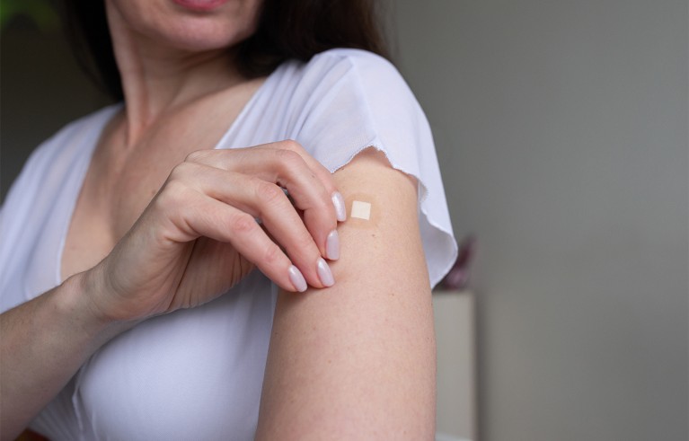 A woman wearing a white top applying a small adhesive hormonal replacement patch on her upper arm.