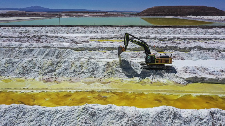 A digger is shown excavating the white lithium salts into neat lines. Bright coloured brine pools are visible in the foreground and background.