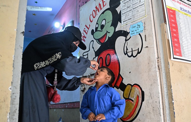 A woman in a black niqab administering oral medicine to a young boy in a blue school uniform inside a classroom. A large, colourful mural of Mickey Mouse with a "WELCOME" sign is painted on the wall behind them, alongside educational posters and charts.