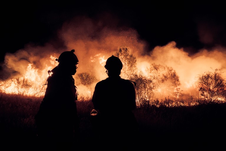 A silhouette of two firefighters looking towards a raging wildfire at night