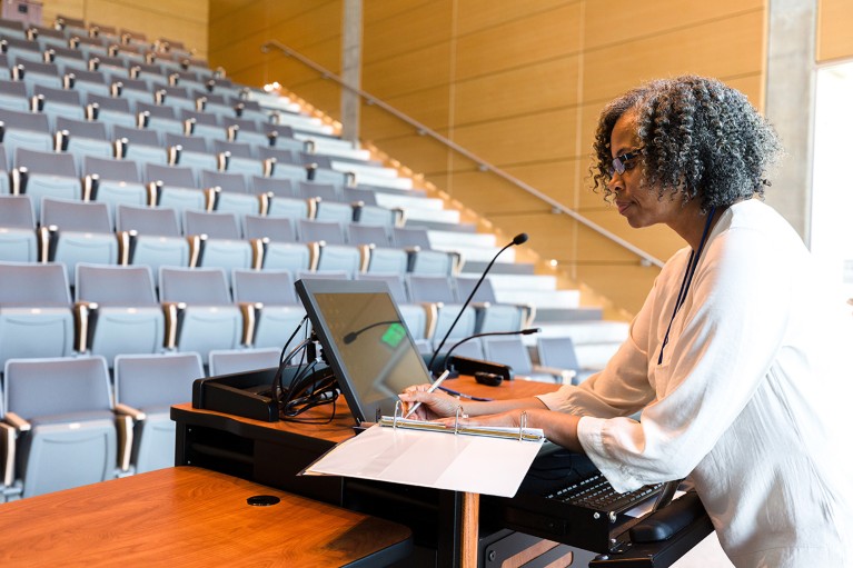 Mature female professor, preparing lecture notes at the lectern, in an empty lecture theatre.