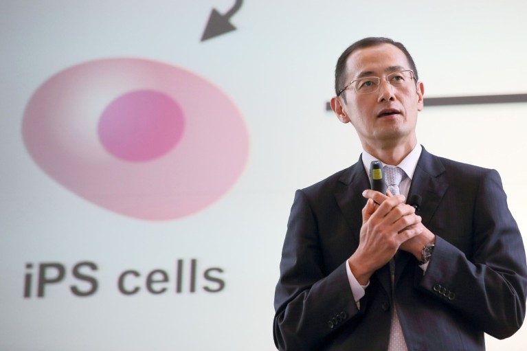 Shinya Yamanaka gives a talk in front of a screen displaying an iPS cell