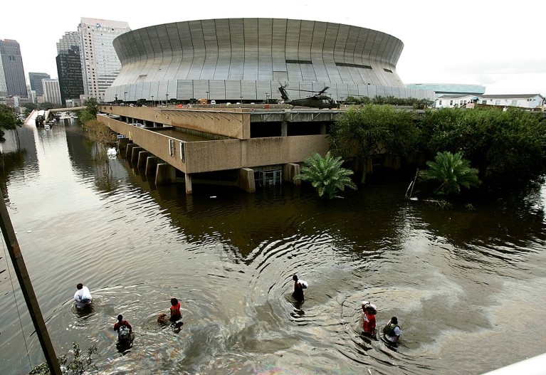 A group of people walk through waist-high water in front of a large stadium.