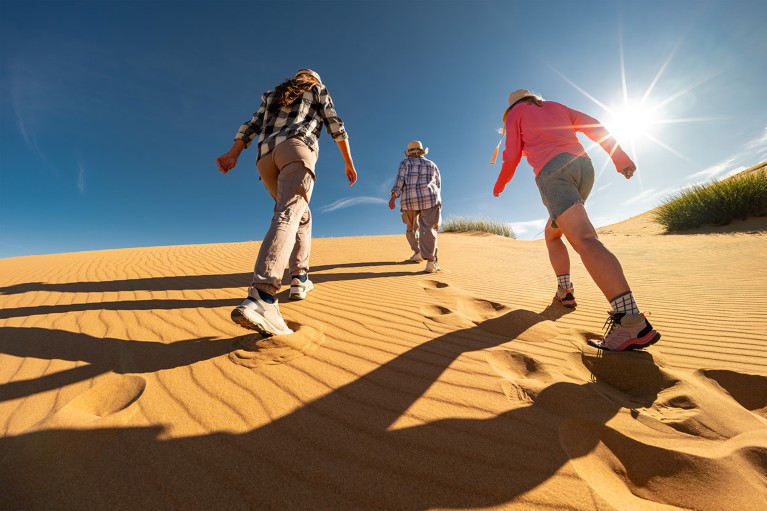 Three hikers climb up a sand dune towards a clear blue sky, leaving footprints in the shifting sand.