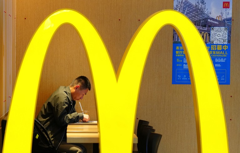 A person sitting and drinking through a straw inside a McDonald's restaurant, framed by a large, illuminated yellow McDonald's arch logo in the foreground. Background features a wooden wall with a blue poster containing text and a QR code, indicating promotional or informational content.