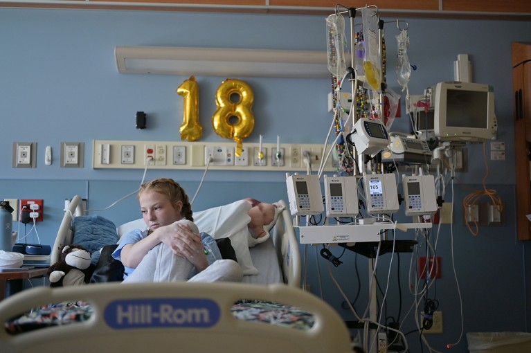 A young woman sits somberly in a hospital bed surrounded by medical equipment. Above her on the wall hangs a bright gold birthday balloon in the shape of the number 18.