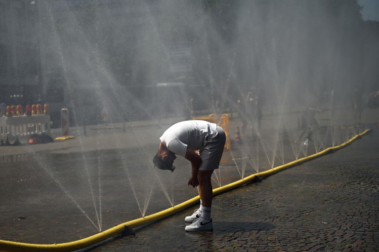 A man bends over to cool his head at a row of water sprinklers in a yellow hose spraying mist across a city street