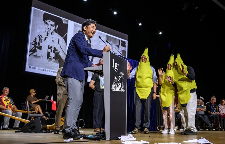 Ig Nobel award ceremony featuring a man speaking at a podium with an "Ig" logo, while three people dressed in banana costumes stand beside him waving. Audience members are seated in the background, and a large black-and-white photo is projected on a screen behind the speaker.