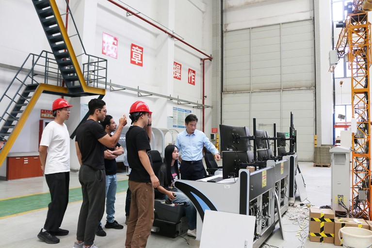 Prof. Dr. Tong Guo points to a panel of computer screens as he instructs a group of five students how to use the smart construction system at Southeast University in Nanjing. The class stands in a large white warehouse space with stairs running up the wall behind them.