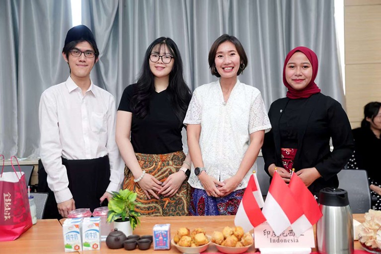 Chairina Hanum stands behind a table with three other Indonesian students at The Business School of Sichuan University's tea event. On the table there are three small red and white Indonesian flags, bowls of snacks and a small black tea pot with four small tea cups.  