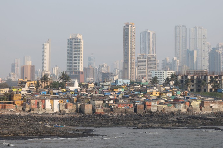 Low-rise slum homes along Mumbai's rocky waterfront, with modern skyscrapers visible in the background on a hazy day.