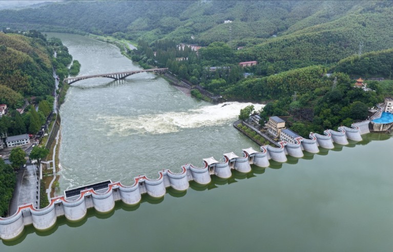 A large dam on a river surrounded by green hills and woodlands, with water flowing through a spillway. Key features include a curved concrete structure with multiple gates, a downstream bridge, and buildings near the dam.